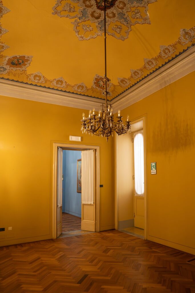 Sala gialla di Palazzo Le Stanze a Pistoia con soffitto decorato, lampadario e porte aperte.