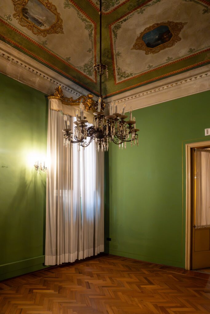 Sala verde di Palazzo Le Stanze a Pistoia con lampadario e tende bianche.