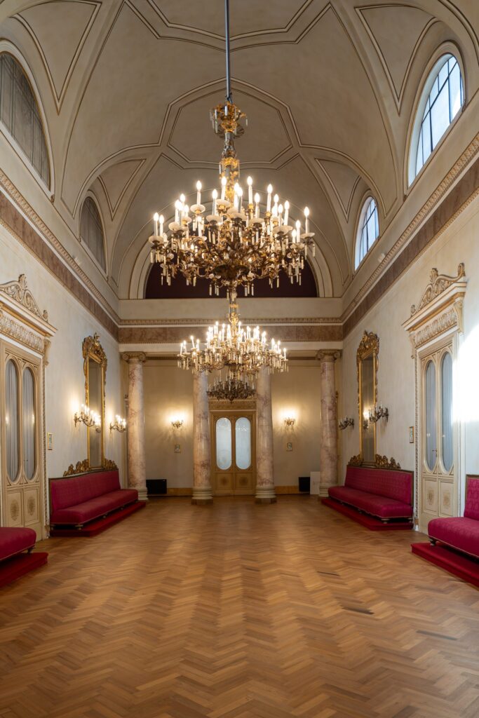 Il Salone delle Feste di Palazzo Le Stanze a Pistoia, con lampadari in cristallo, divani rossi e parquet.